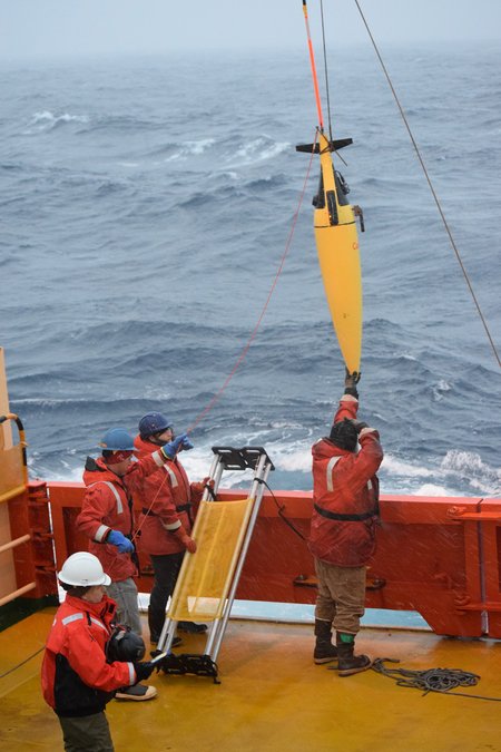 People in bright orange gear stand on a boat in the sea. A yellow oblong shape extends from a rope out of frame.