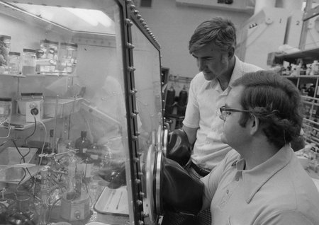 Two men looking into the chemistry hood while the man in the foreground works with a flask in the hood. The older man in the background is Fred Anson.