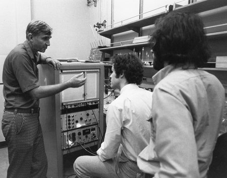 A man is describing something to two other men, standing next to a piece of lab equipment.