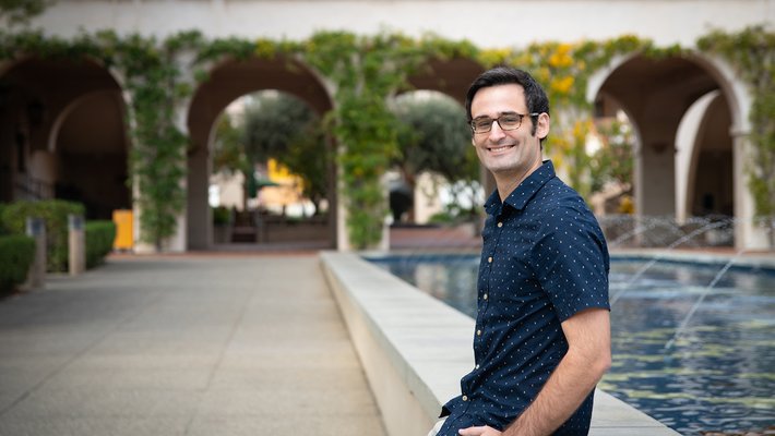 A portrait of Peter Caradonna. He wears a short-sleeve button up shirt and slacks. He sits casually in front of a fountain, smiling.