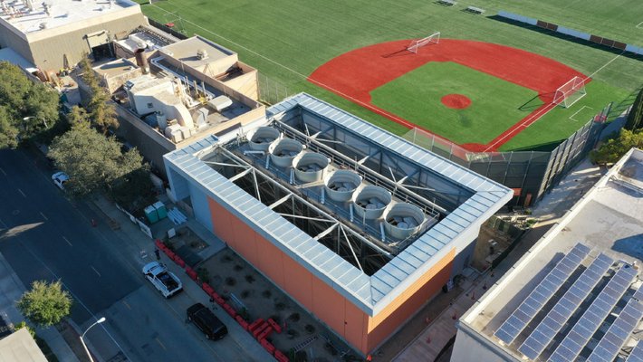 Aerial view shows a baseball diamond adjacent to four buildings with equipment or solar panels on their rooftops. Six cylindrical towers jut out of the central building, each with a metal fan visible inside it. In the foreground, trees, a few cars, and orange construction barriers line a paved, two-lane street.