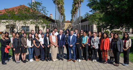 A group of scholars stand on a walkway in front of two rows of palm trees with Stanford buildings visible in the background.