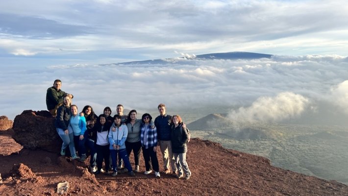 Sunlight illuminates the faces of a dozen people standing close together on an ochre ground of soil and rocks that abruptly ends, giving way to a view of a green plain far below dotted with cinder cones, a large mountain in the distance, and interwoven clouds and clear sky.