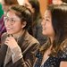 Two women with brown hair are shown in profile, smiling presumably at a speaker during a session at a symposium.