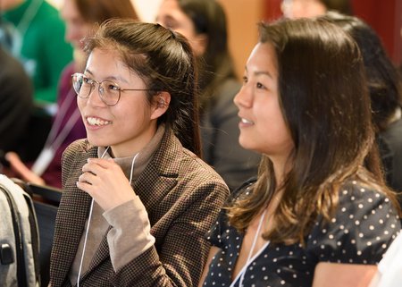 Two women with brown hair are shown in profile, smiling presumably at a speaker during a session at a symposium.