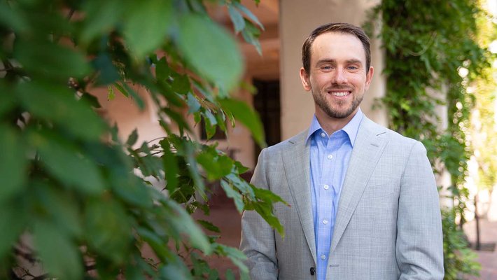 A portrait of Pawel Janas. He stands outside smiling and wearing a suit. There are columns with vines behind him.