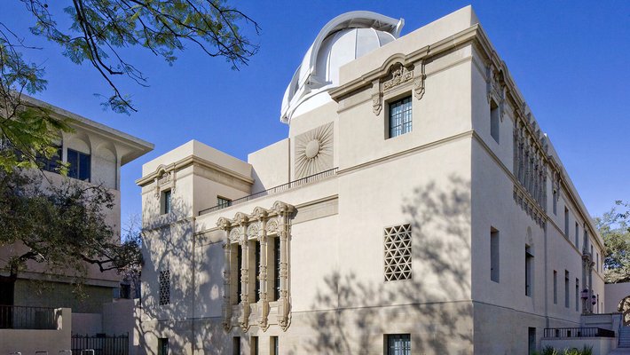 A cream-colored, three-story building with ornate sculptures including a bas relief of the sun behind the broad, third-story balcony. An open, white telescope dome rises from the flat roof of the building, with a bright blue sky above. In the foreground is a decomposed-granite landscape and a few tree branches, and on the left a slightly taller concrete buiding.
