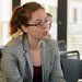 Close up photo of a. woman with red hair, glasses, and a plaid blazer, appearing to be talking during a session at a symposium.