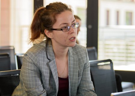 Close up photo of a. woman with red hair, glasses, and a plaid blazer, appearing to be talking during a session at a symposium.