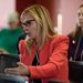 A close up photo of a blond woman in an orange jacket and glasses talking from her seat at a symposium.