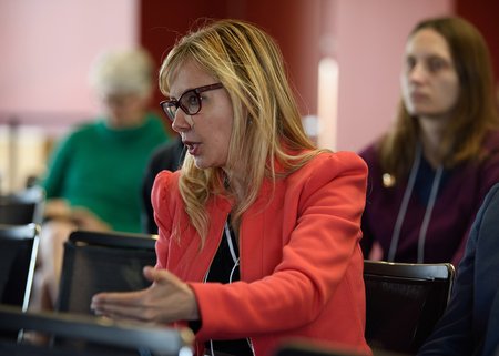 A close up photo of a blond woman in an orange jacket and glasses talking from her seat at a symposium.