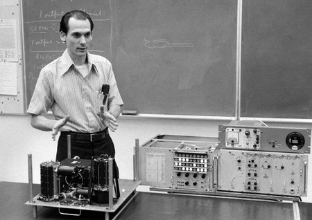 A man standing behind a physics experiment demonstration table in front of a blackboard